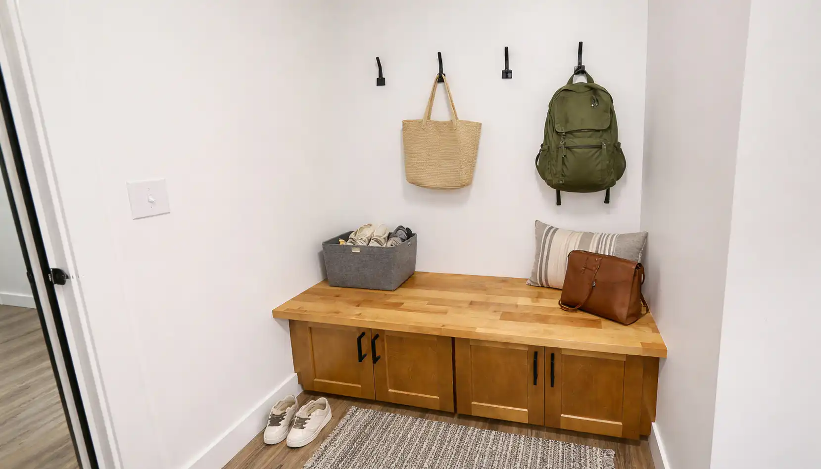 Leo mudroom storage bench with wood cabinets, black hooks, bags, shoe bin, and striped pillow.